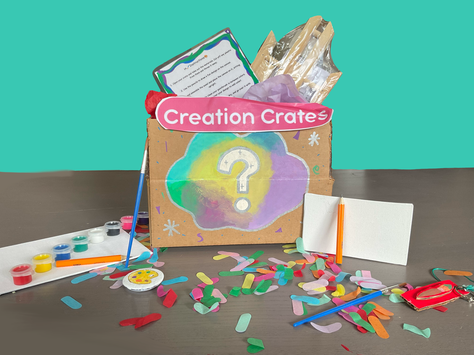 A box with the creation crate logo on it. an easel and instruction manuel is peaking out of the box. art suplies and confetti are on the table.